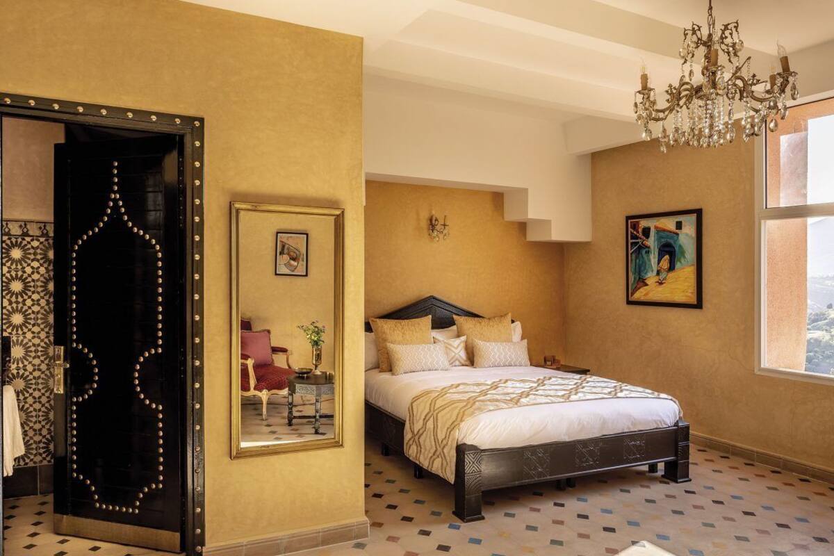 A bedroom with a bed, a chandelier, a mirror, and a window.