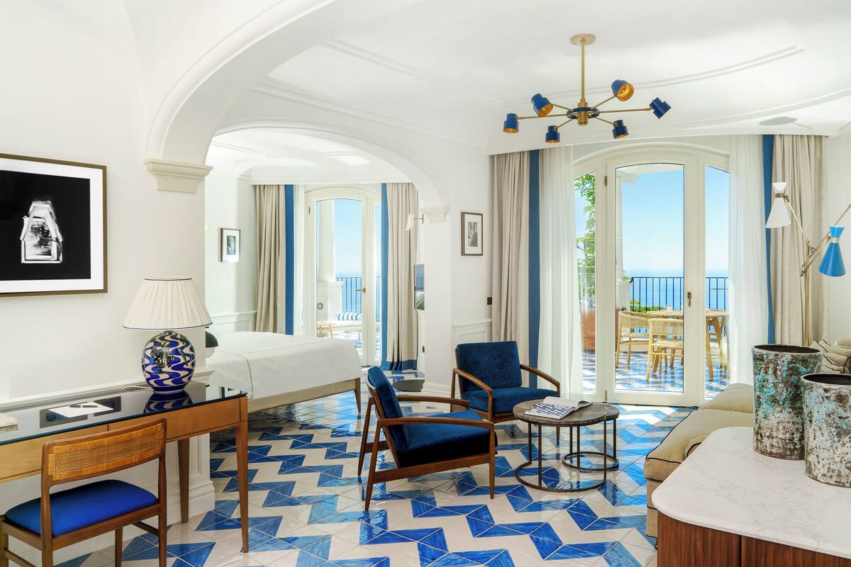 Hotelkamer met een bed, twee blauwe fauteuils, een bureau met een lamp en uitzicht op het omliggende landschap door grote ramen.