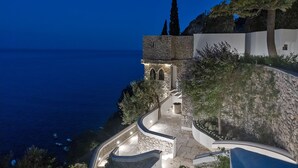A stone pathway leading to a building with a stone facade and arched windows, overlooking a coastline. - Borgo Santandrea (Amalfi)