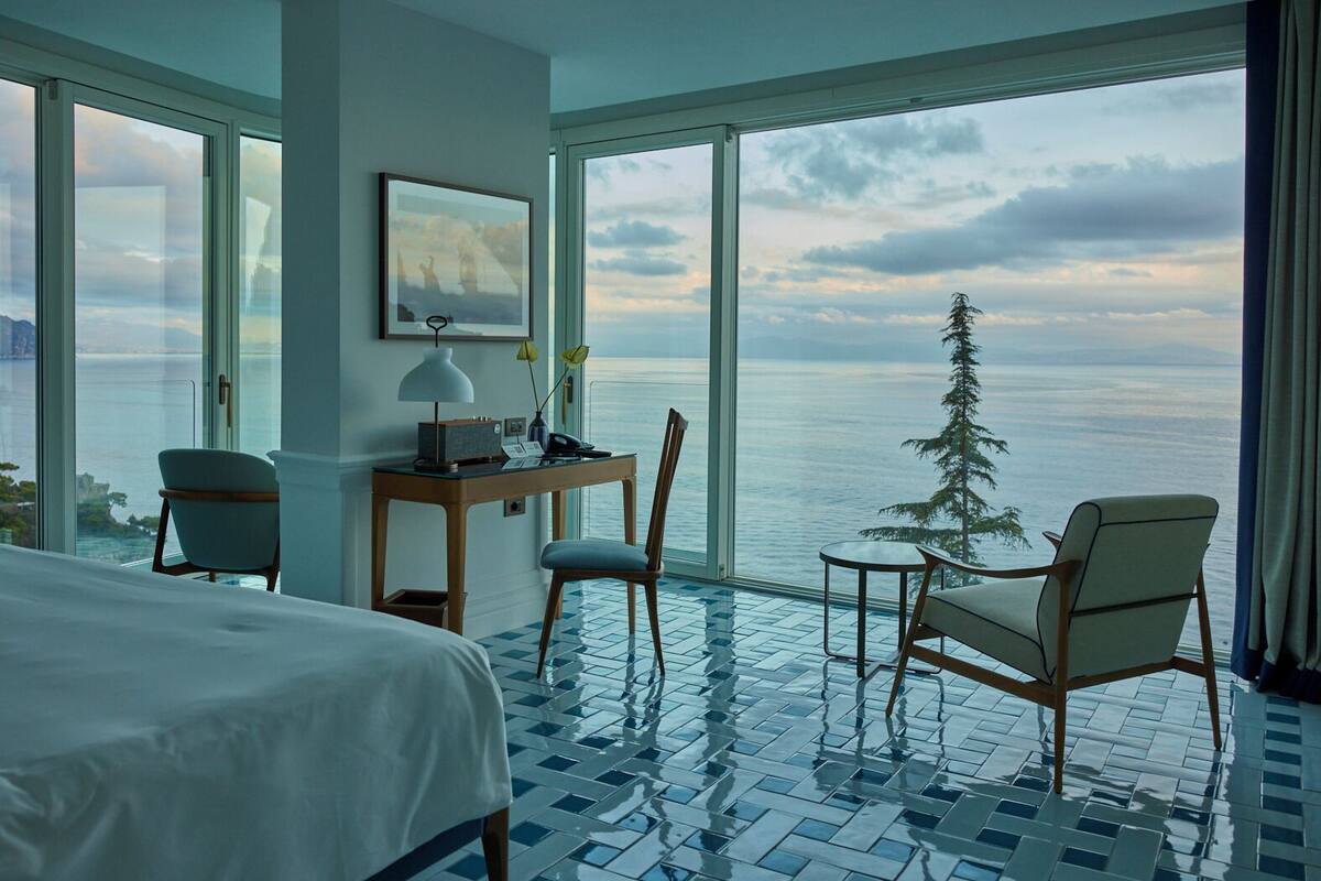 Hotelkamer met groot raam met uitzicht op zee en een boom, een bed, bureau met lamp en twee stoelen.