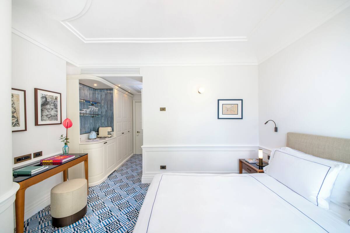 Een hotelkamer met een groot bed, een nachtkastje met een lamp, een tafeltje met boeken, een decoratieve vaas en uitzicht op de badkamer.