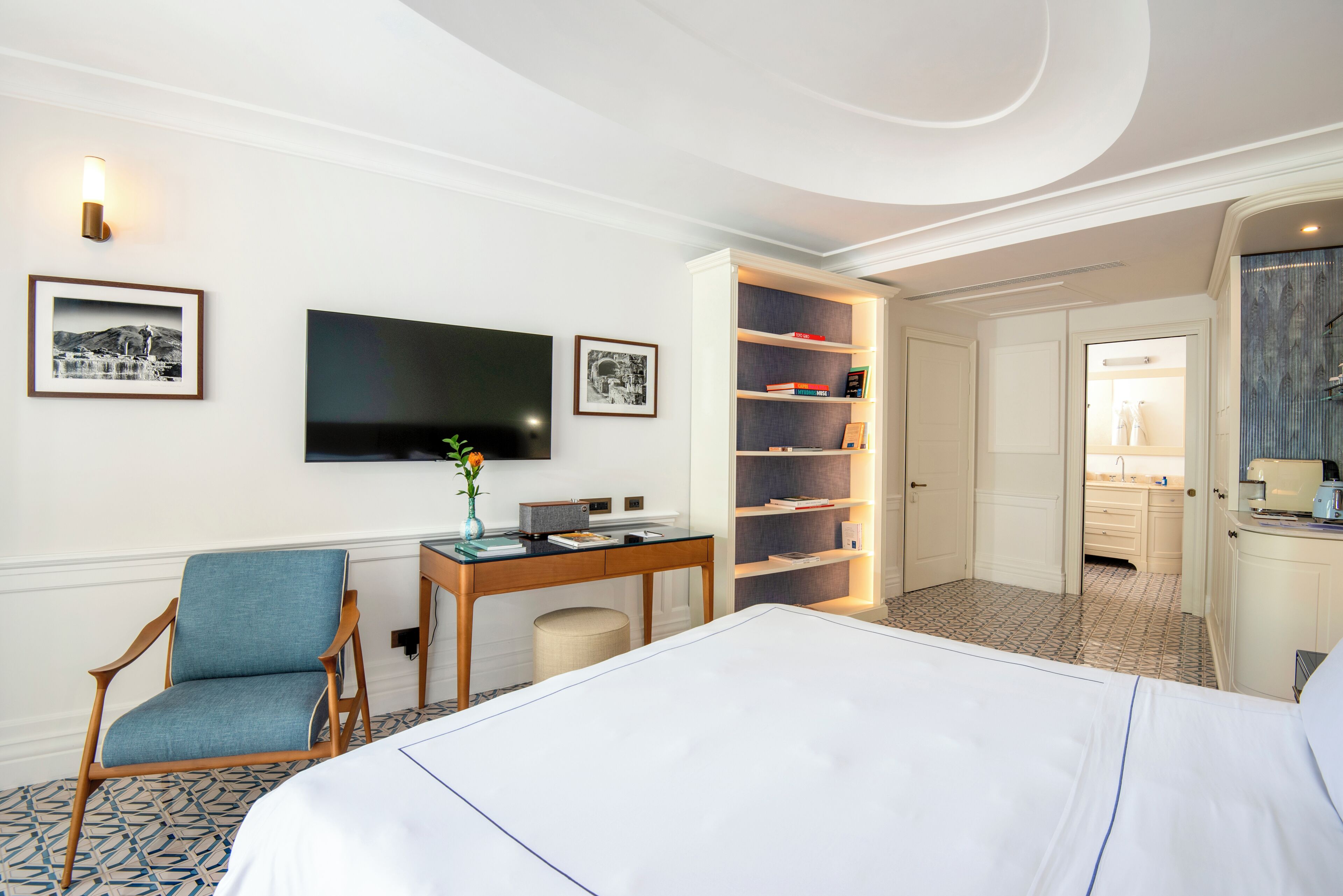Habitación de hotel con cama, escritorio, silla, televisor, estantería y baño visible a través de una puerta abierta.