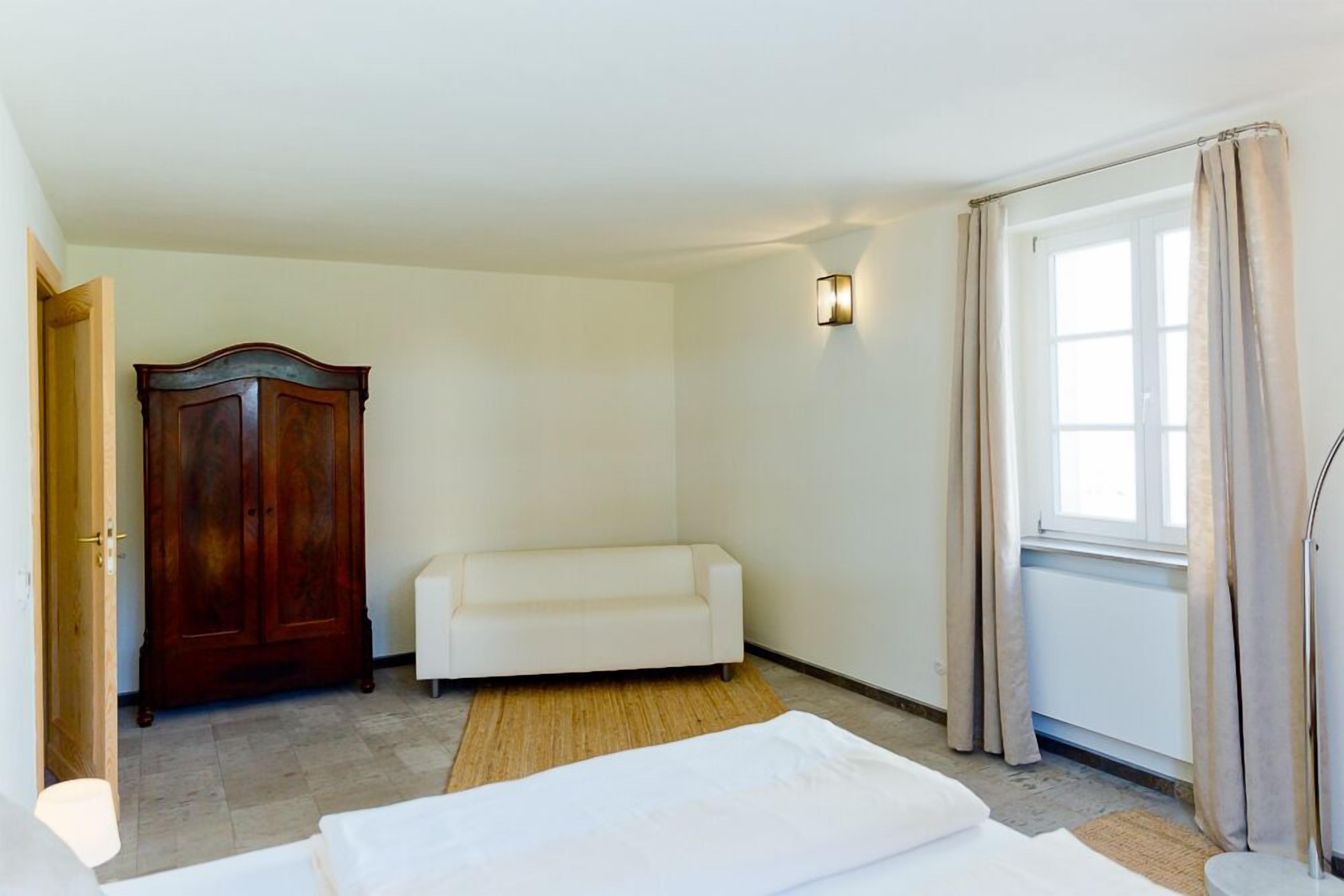 A hotel room with a wooden wardrobe, a white sofa, a bed with white bedding, a wall-mounted light fixture, and a window with curtains.