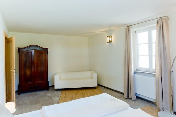 A hotel room with a wooden wardrobe, a white sofa, a bed with white bedding, a wall-mounted light fixture, and a window with curtains. - Gästehaus am Lehnitzsee (Potsdam)