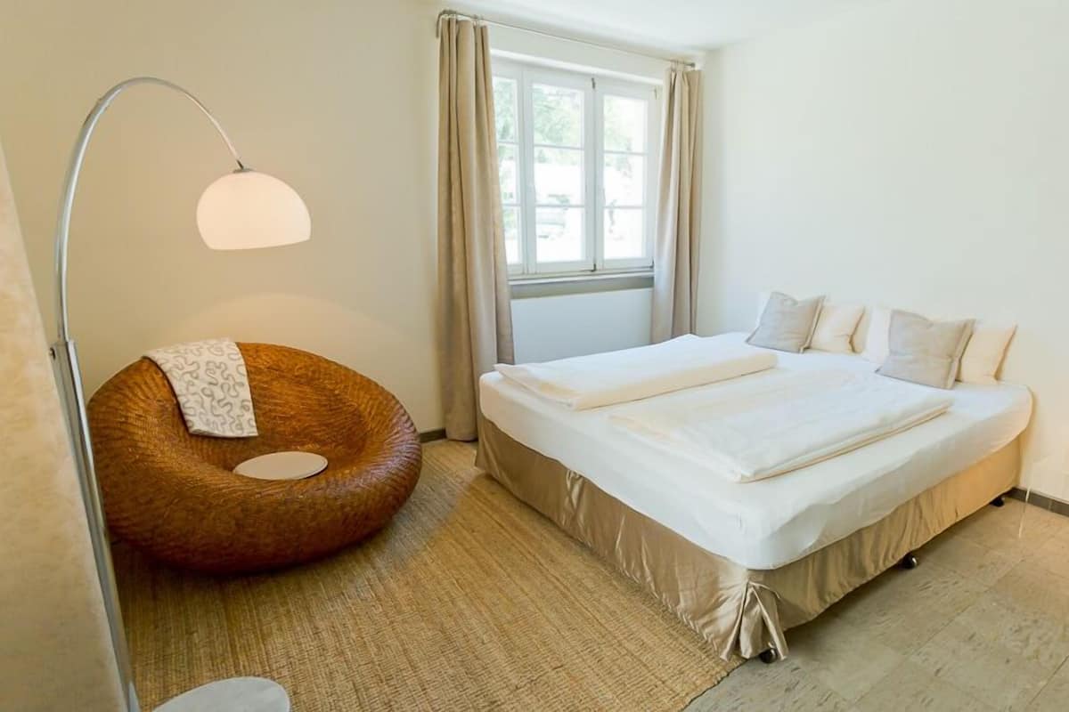 Ein modernes Schlafzimmer mit einem großen Bett, einem runden Sessel, einer Stehlampe und einem Fenster mit Vorhängen.