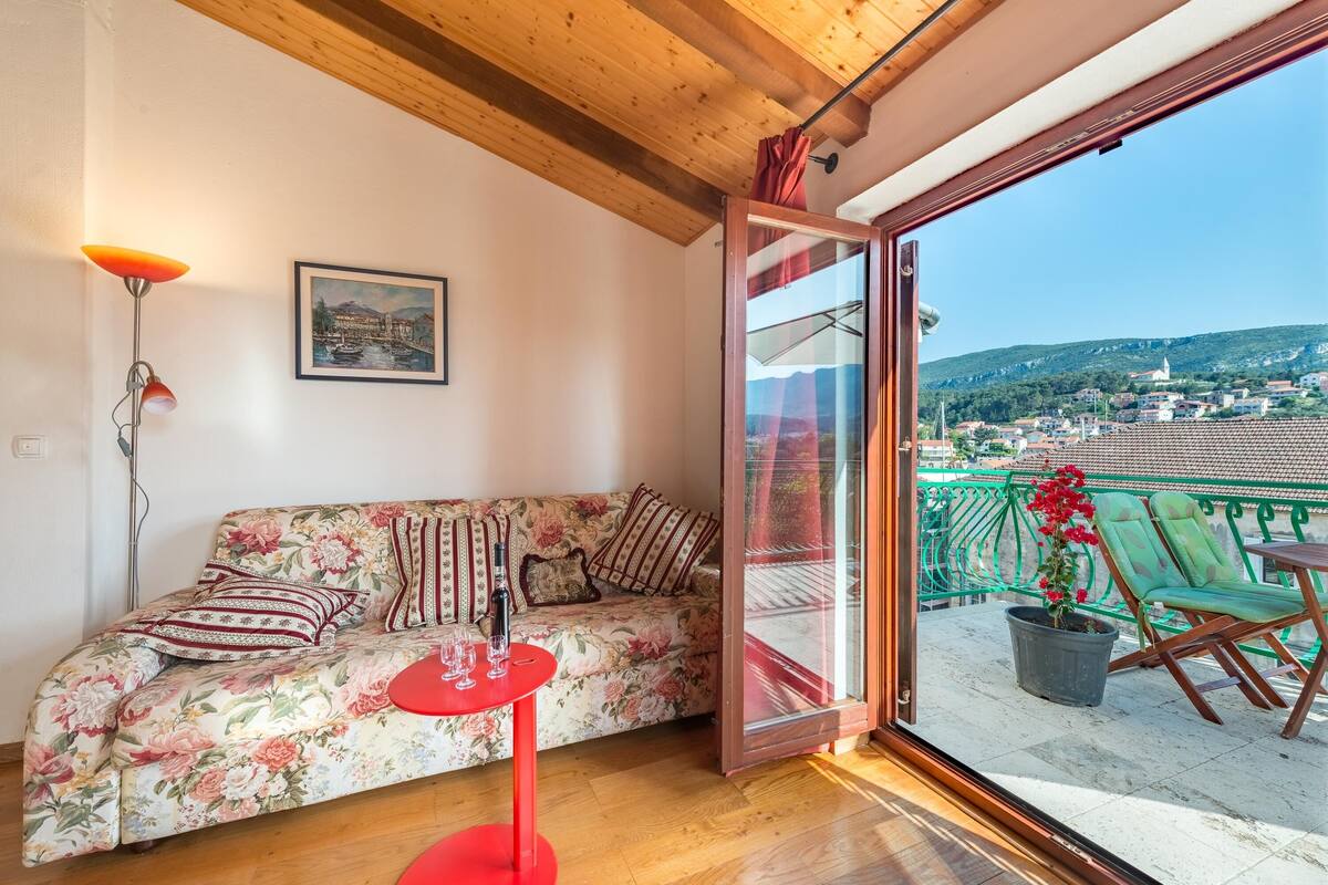 Um quarto aconchegante com um sofá floral, uma mesa vermelha e uma varanda com vista para uma cidade e montanhas.