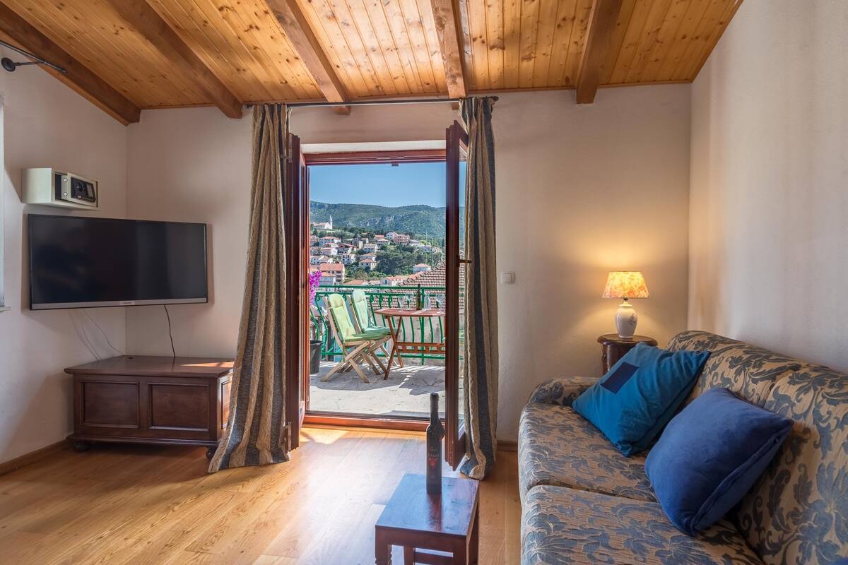 Quarto com teto de madeira, televisão, sofá com almofadas azuis, mesa pequena e vista para uma varanda com cadeiras e mesa.