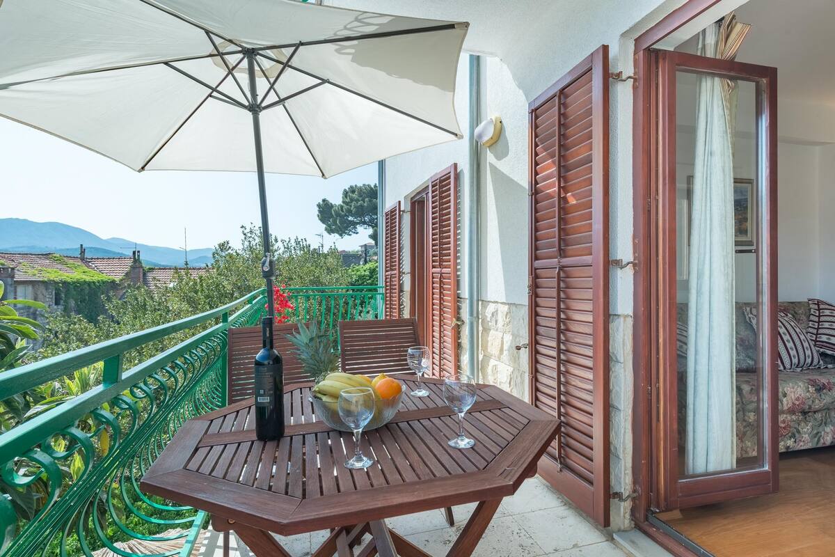 Varanda com mesa de madeira, guarda-sol branco e uma tigela com frutas e copos de vinho.
