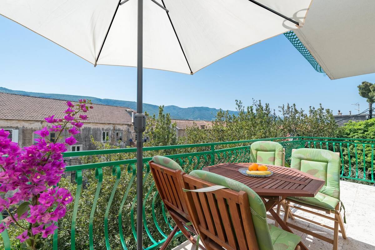 Varanda com mesa e cadeiras de madeira, um guarda-sol branco e vista para edifícios e montanhas.