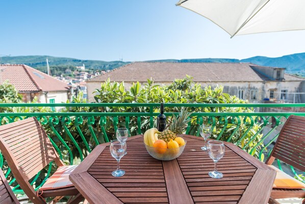 Une table en bois, sur laquelle se trouvent un panier de fruits et des verres à vin, est placée sur un balcon doté d’une rambarde verte, d’où l’on peut admirer une ville avec ses bâtiments et ses collines.