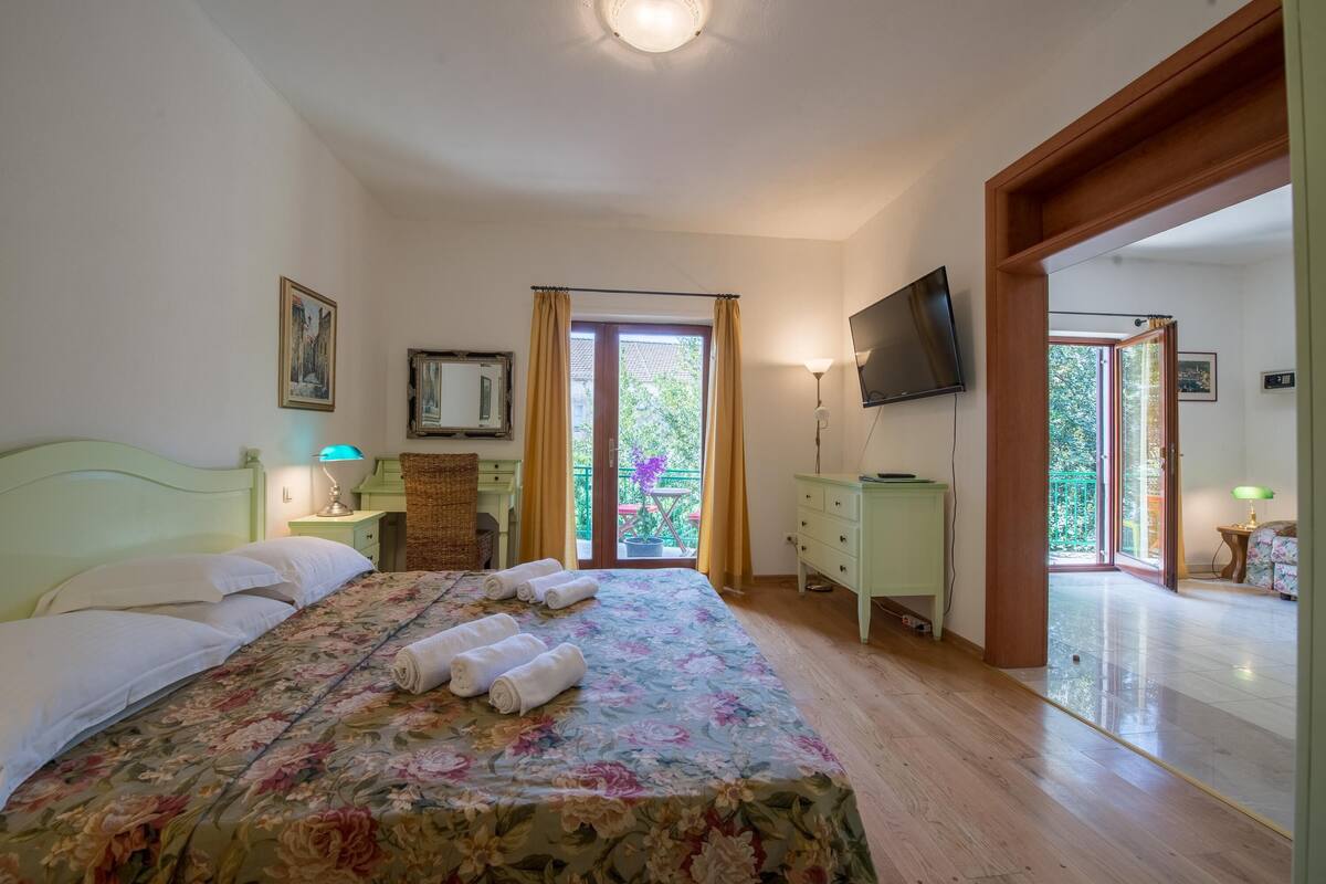 Quarto com cama, televisão, escrivaninha e varanda com cortinas.