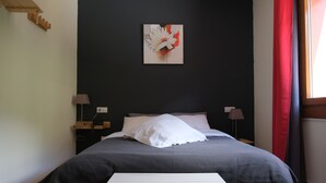 Ein ordentlich gemachtes Bett mit grauer Decke, weißen Kissen und einem kleinen Nachttisch. Darüber hängt ein Wandbild und zur Rechten befindet sich ein Fenster mit Vorhängen.