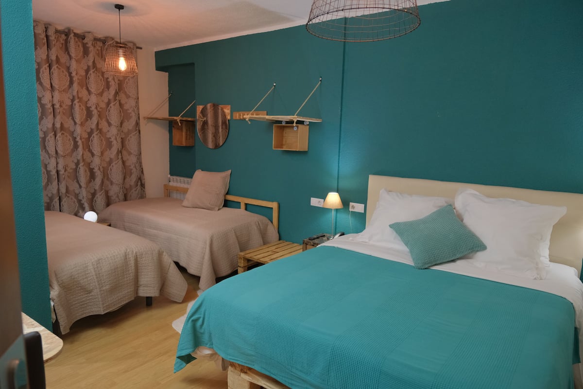 A room with two single beds, a larger bed, teal walls, wooden furniture, and a hanging light fixture.