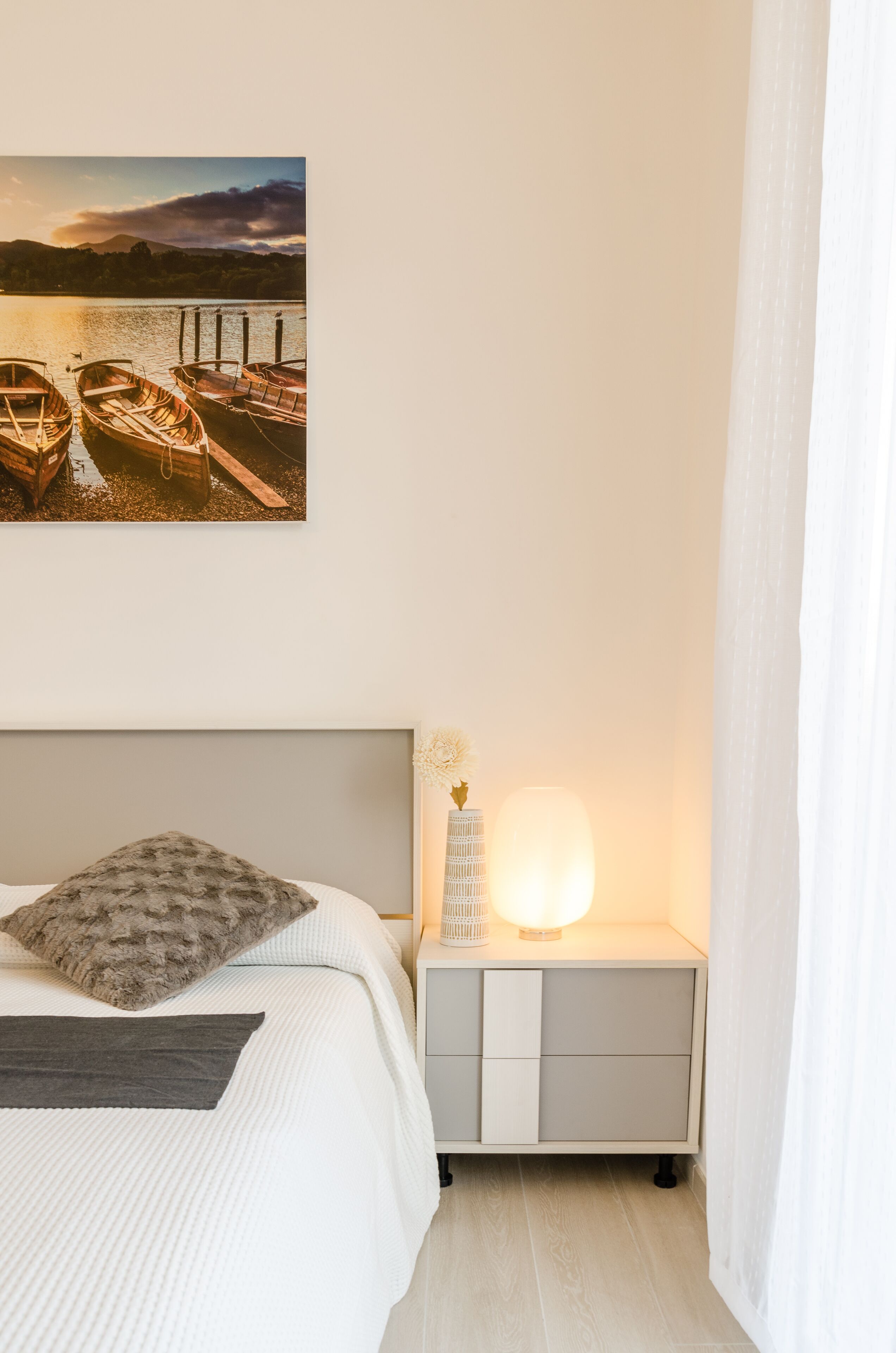 A bedroom with a bed, a bedside table with a lamp, a nightstand, and a picture of boats on the wall.