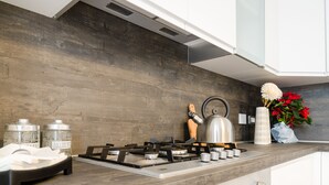 Une cuisine moderne avec un revêtement mural en bois, une cuisinière à gaz et une bouilloire. On y trouve deux récipients étiquetés « ZUCCHERO » et un vase avec des fleurs rouges.