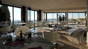 A modern dining area with round tables and white chairs, large windows offering a view of the cityscape. - Hotel La Montagne de Brancion (Martailly-Les-Brancion)