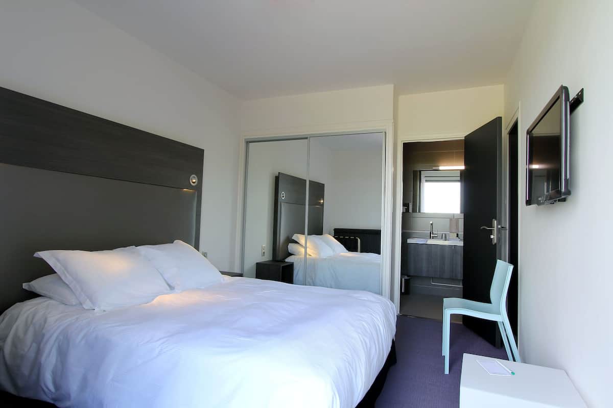 Une chambre d’hôtel avec deux lits, une chaise, un bureau et une salle de bain visible à travers une porte ouverte.
