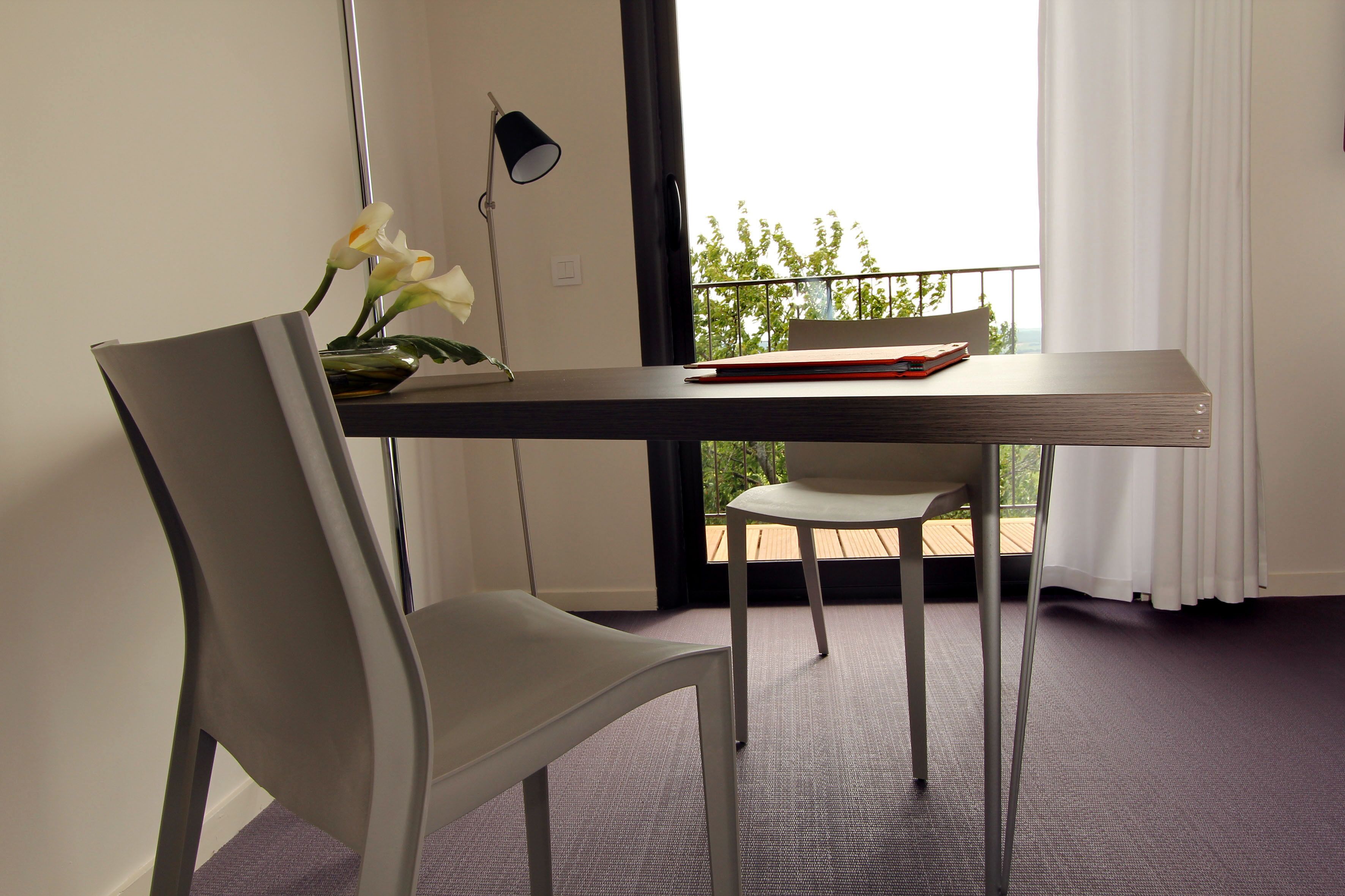 A modern room with a dining table, two chairs, a vase with flowers, a lamp, and a view of greenery outside.