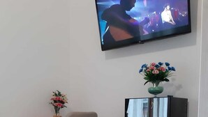 A modern living room with a flat-screen TV mounted on the wall, a white desk with a chair, and a small shelf with a vase of flowers. - English Inn rooms (Sorrento)