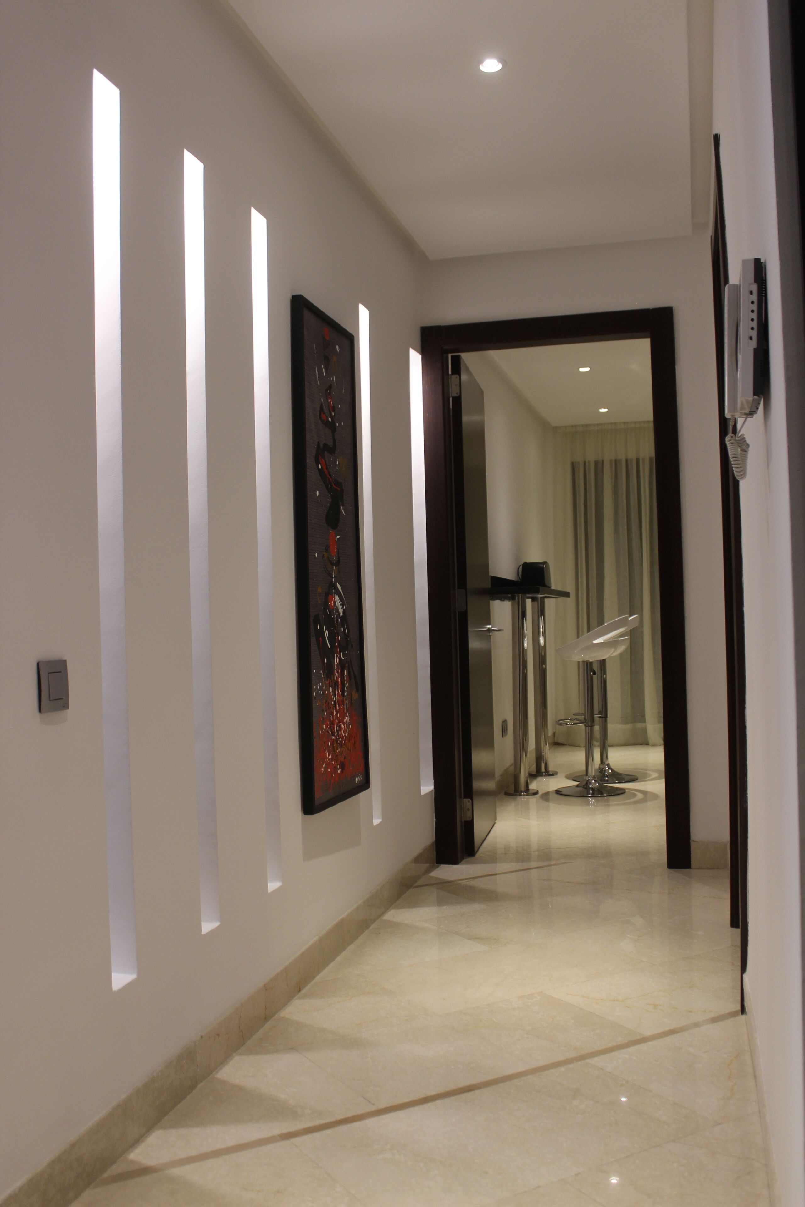 A modern hallway with recessed lighting, a framed artwork, and a glass door leading to a room with a mirror and a chair.
