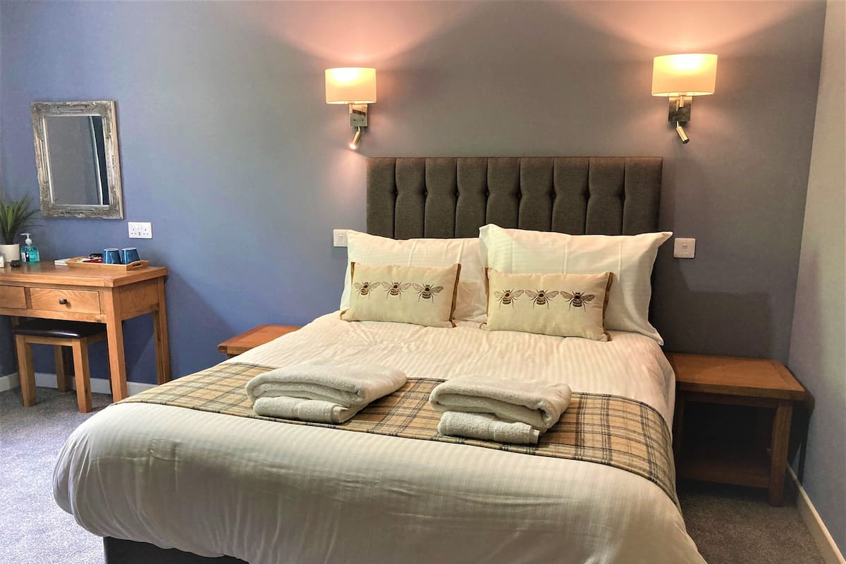A neatly made bed with two pillows, a bedside table, and a desk with a chair.