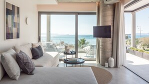 A modern hotel room with a large bed, a TV, a balcony with a view of the sea, and a dining area. - Urban Suites (Alimos)