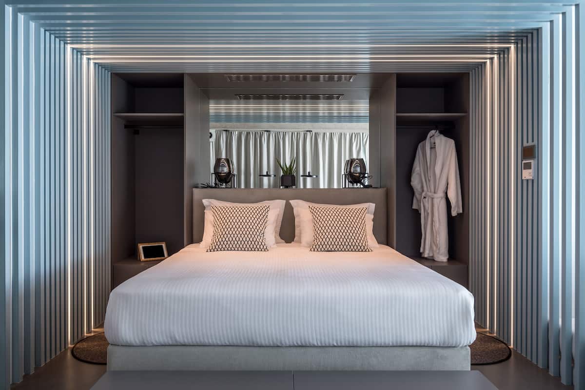 A modern bedroom with a large bed, two bedside tables, a robe hanging on the wall, and a view of the outside through a window with blinds.