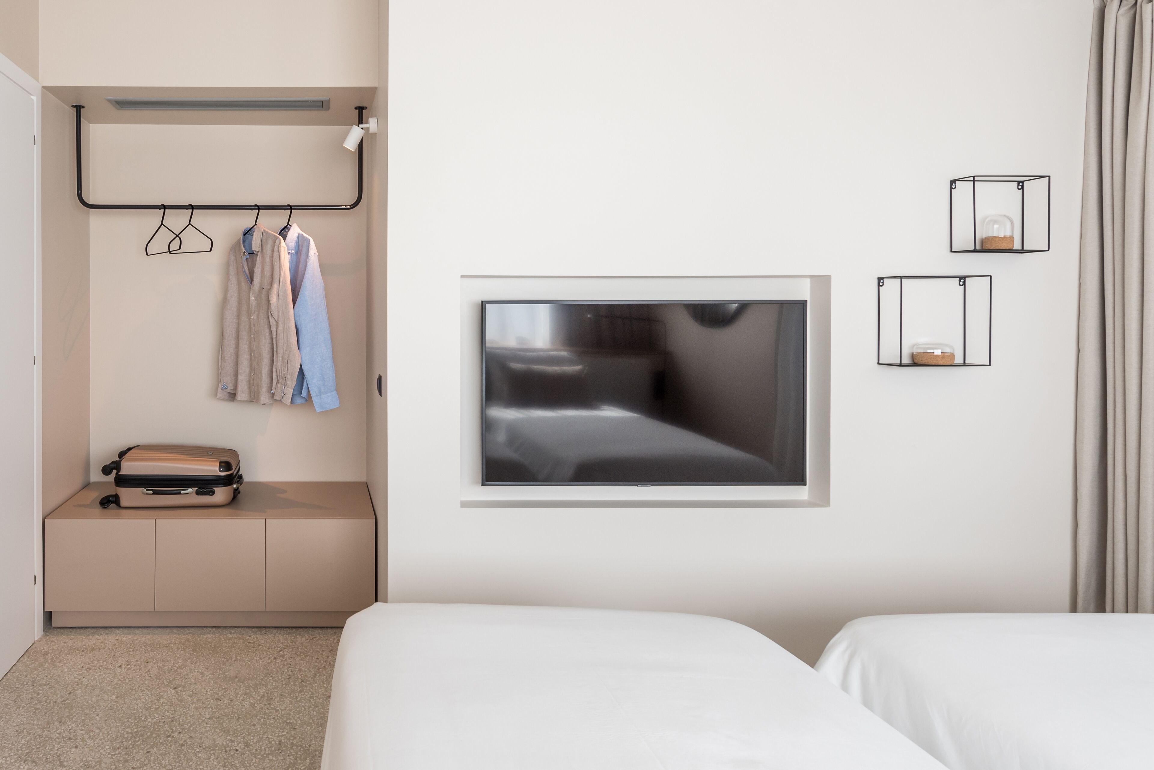 Habitación de hotel con cama, zona de estar con maleta, perchero con ropa y un cuadro en la pared.