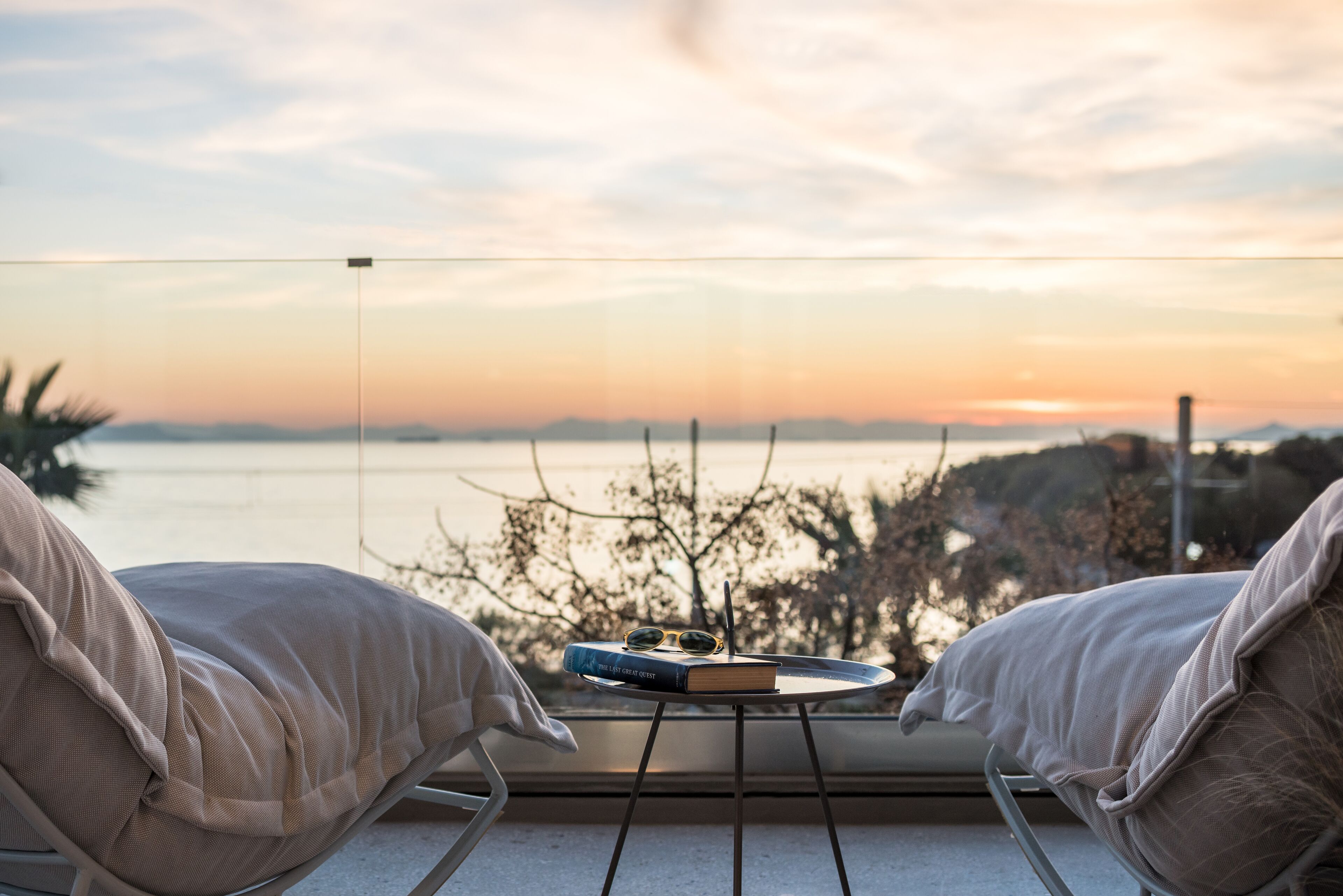 A balcony with two chairs, a table with a book and sunglasses, and a view of the sea at sunset.