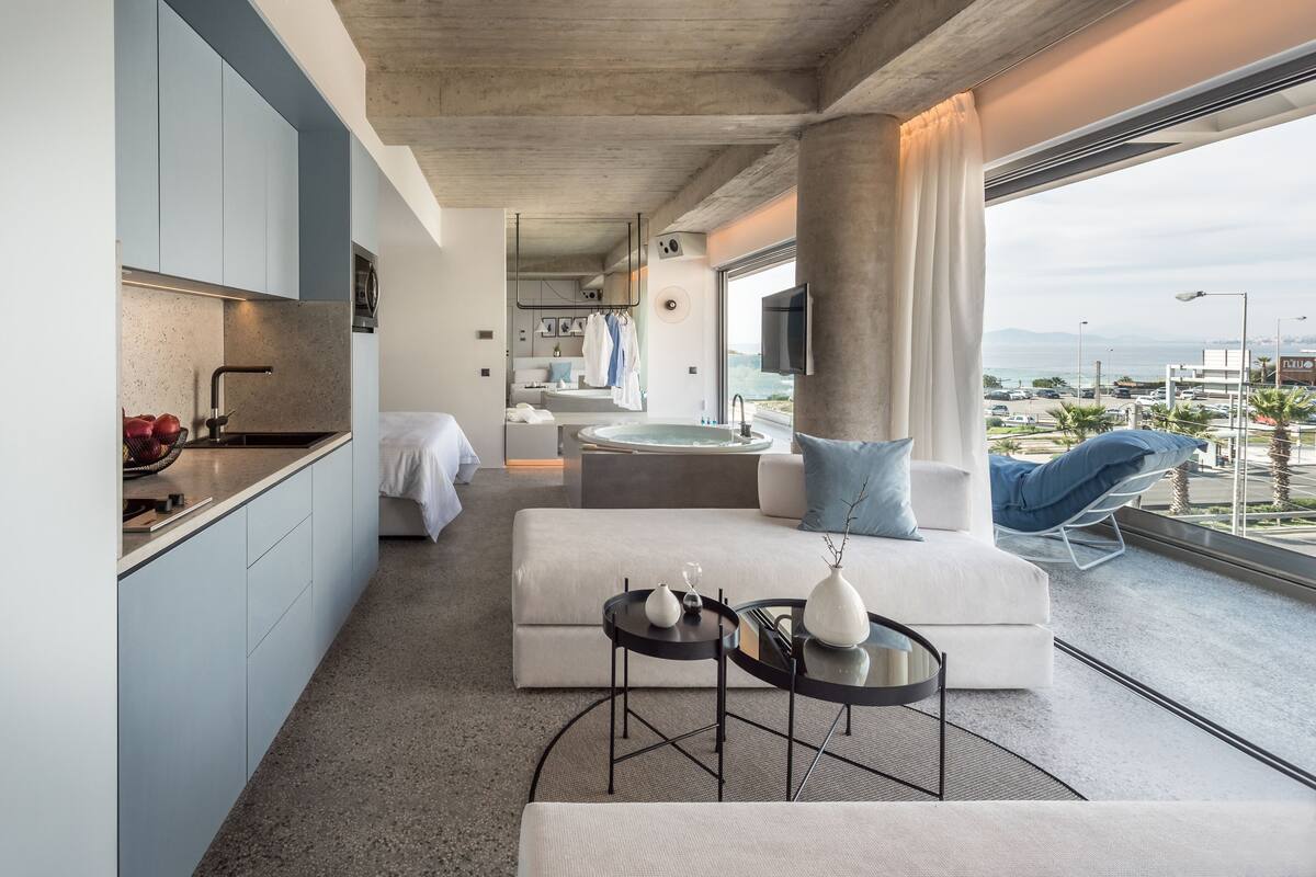 A modern hotel room with a kitchen, a living area with a sofa and coffee table, a balcony with a view, and a large window.