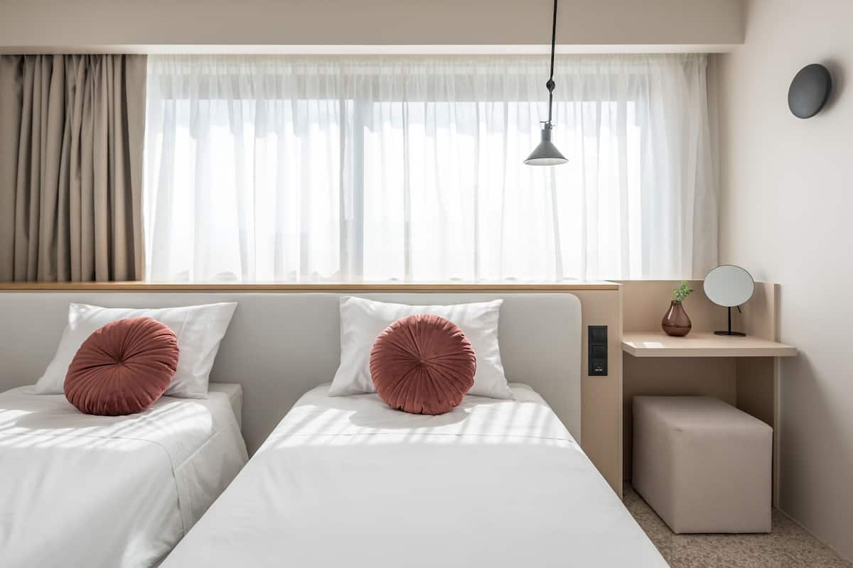 A hotel room with two beds, a bedside table with a mirror, a small stool, and a window with sheer curtains.