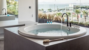 A modern bathroom with a large bathtub, a window with a view of the sea and cityscape, and a minimalist design. - Urban Suites (Alimos)