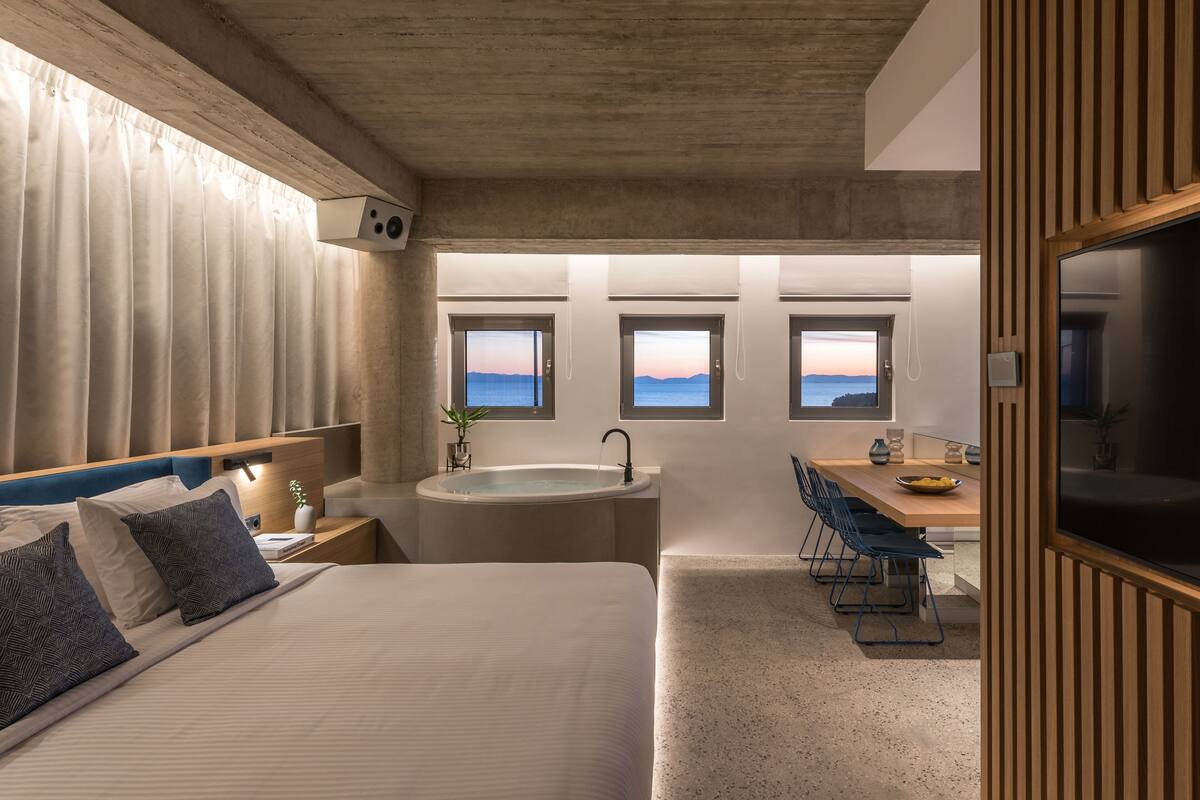 A modern bedroom with a large bed, a built-in desk, and a view of the sea through windows.