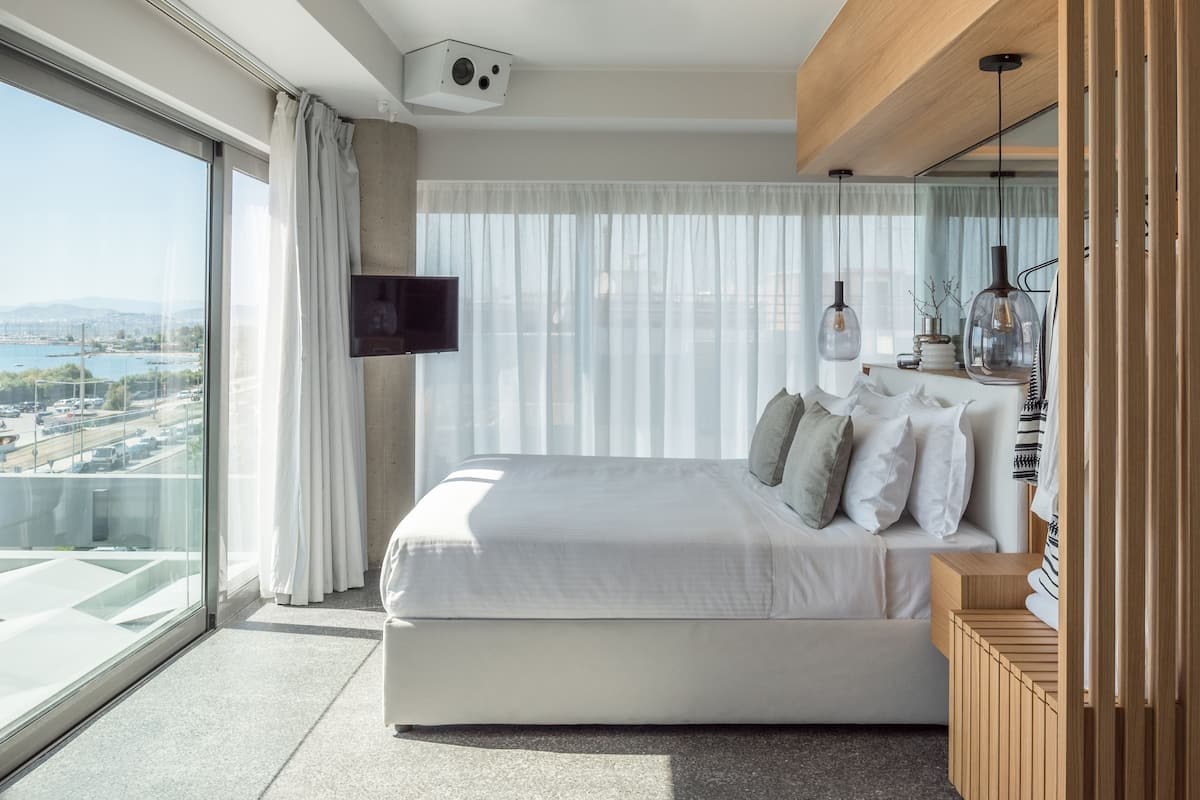 A modern hotel room with a large bed, a flat-screen TV, a wooden bedside table, and a view of a cityscape through floor-to-ceiling windows.