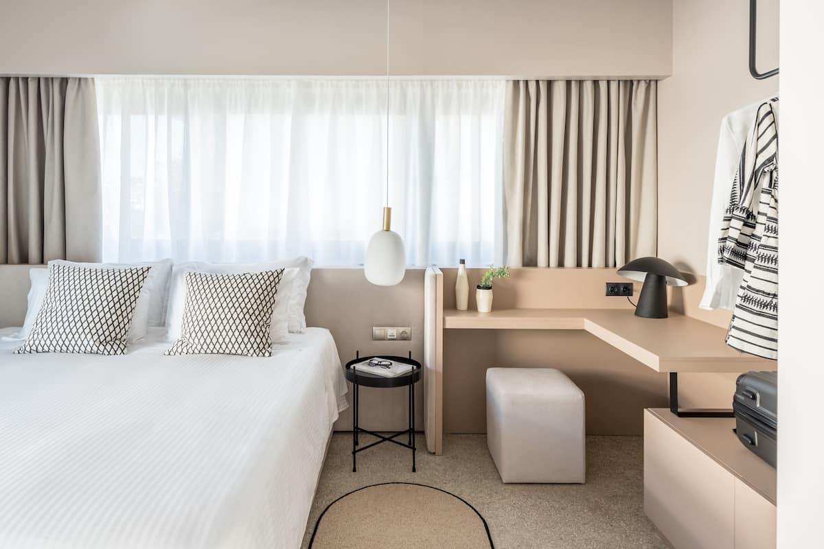 A modern hotel room with a bed, bedside table, desk, and a small seating area.