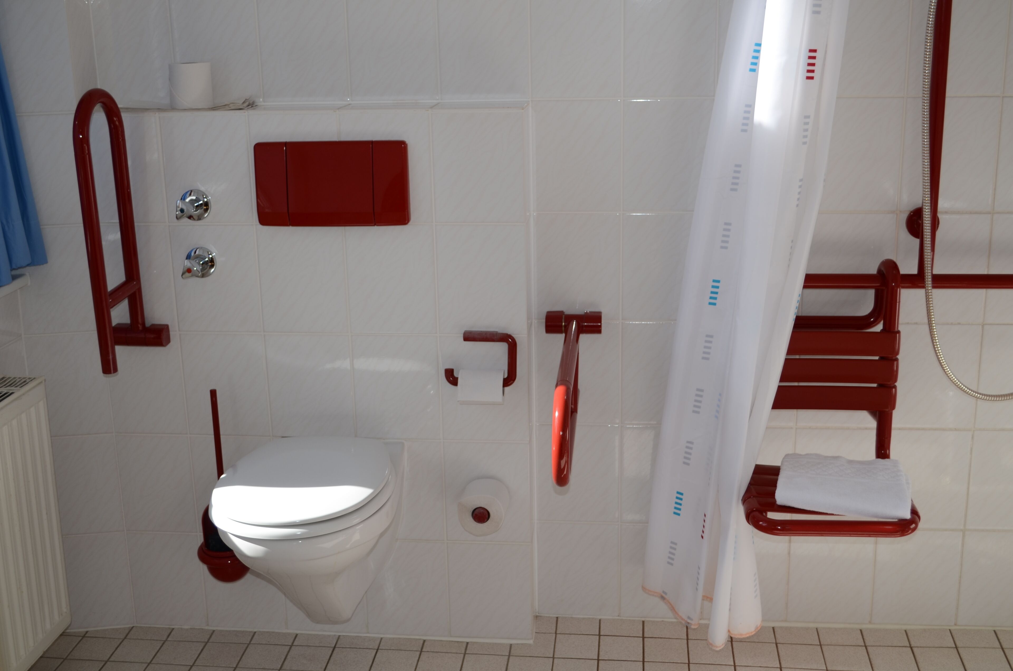 A bathroom with a toilet, a red grab bar, a white shower curtain, and a towel rack.