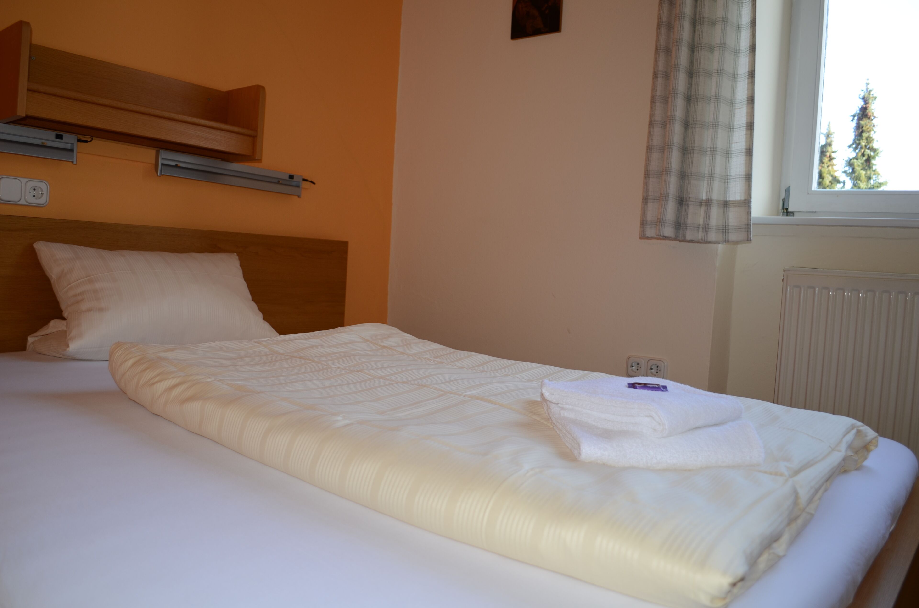 A neatly made bed with a white duvet and a folded towel, a wooden headboard, and a window with a view of trees.