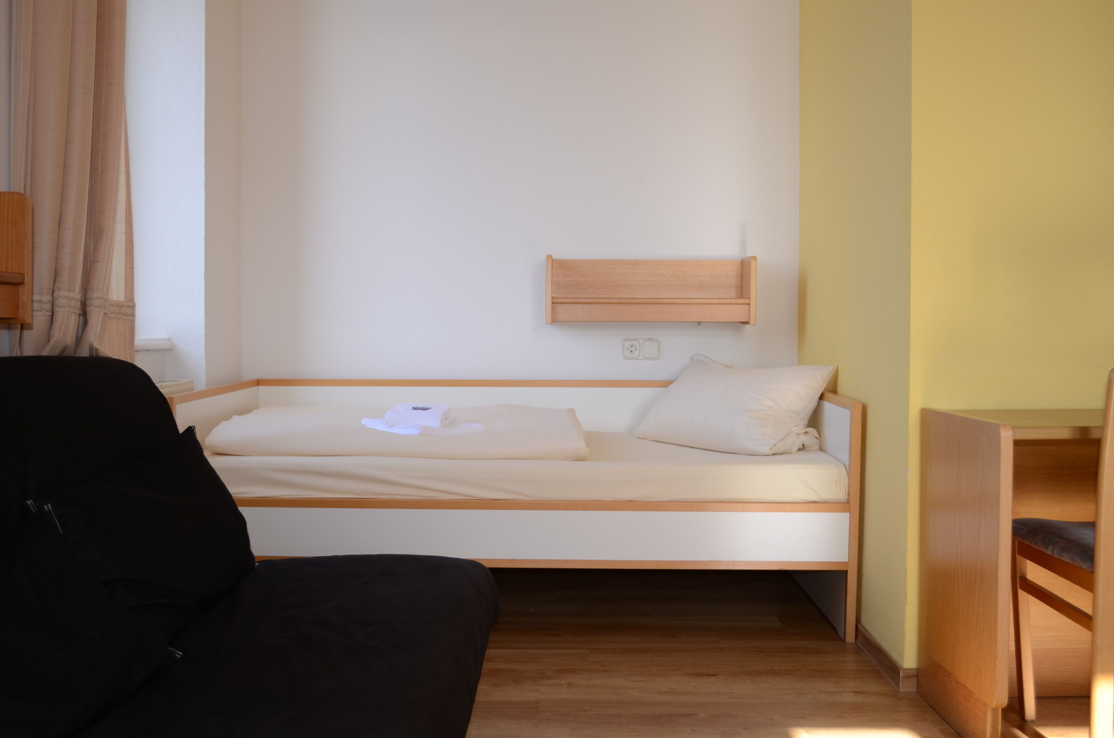 A small, single-bed room with a wooden headboard, a bedside table, and a black chair.