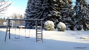 A snow-covered playground with swings and a ladder, surrounded by tall evergreen trees. - Stiftsberg Bildungs und Freizeitzentrum (Kyllburg)
