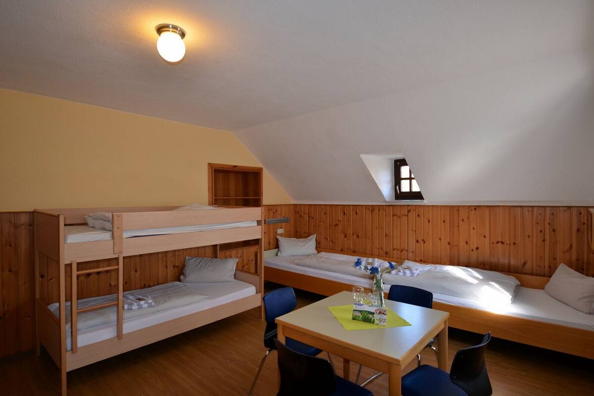 A dormitory room with bunk beds, a table with chairs, and a small window.