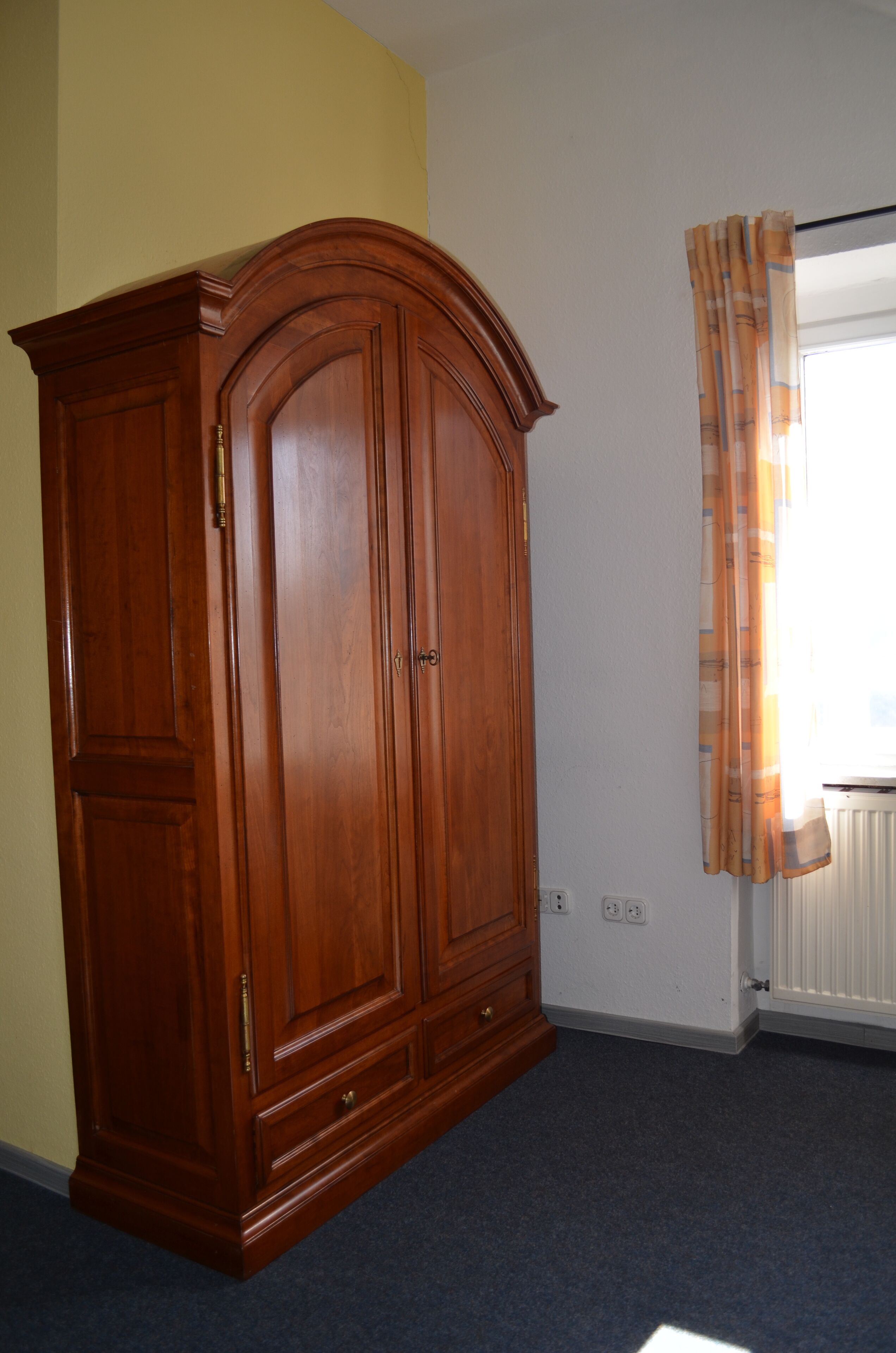A room with a wooden wardrobe, a radiator, and a window with curtains.