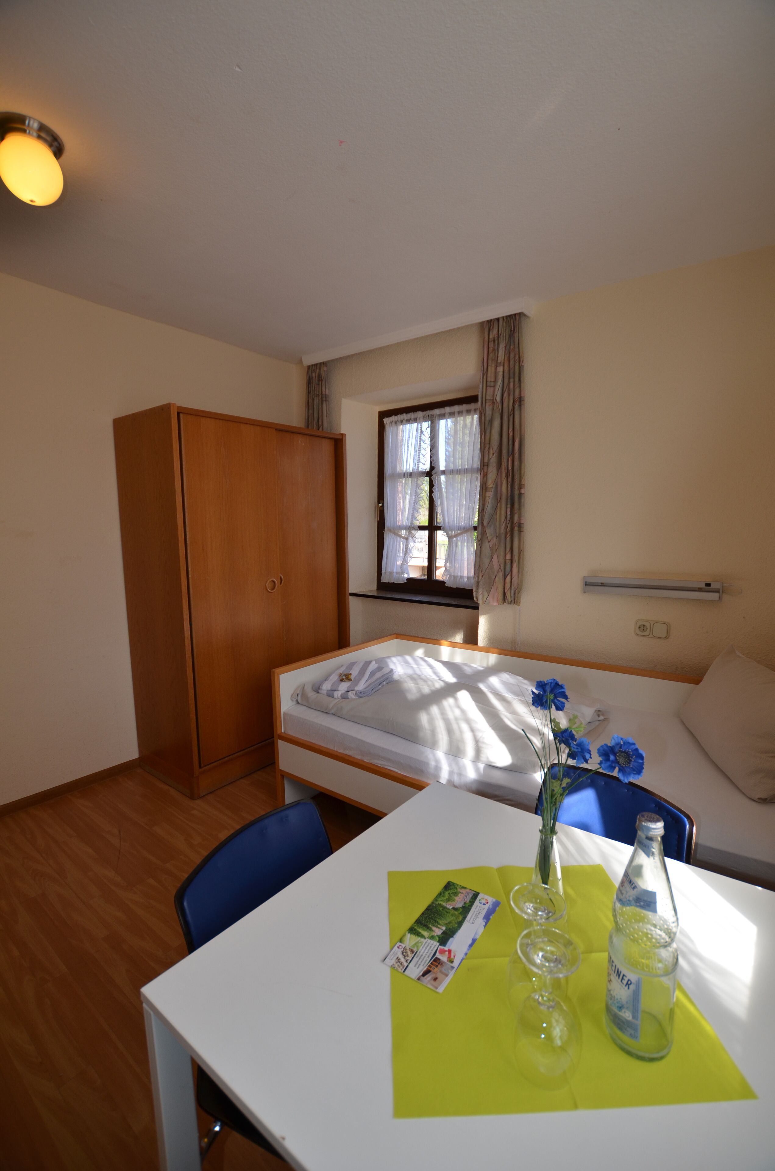 A hotel room with a bed, a wardrobe, a table with a bottle and glasses, and a window with curtains.