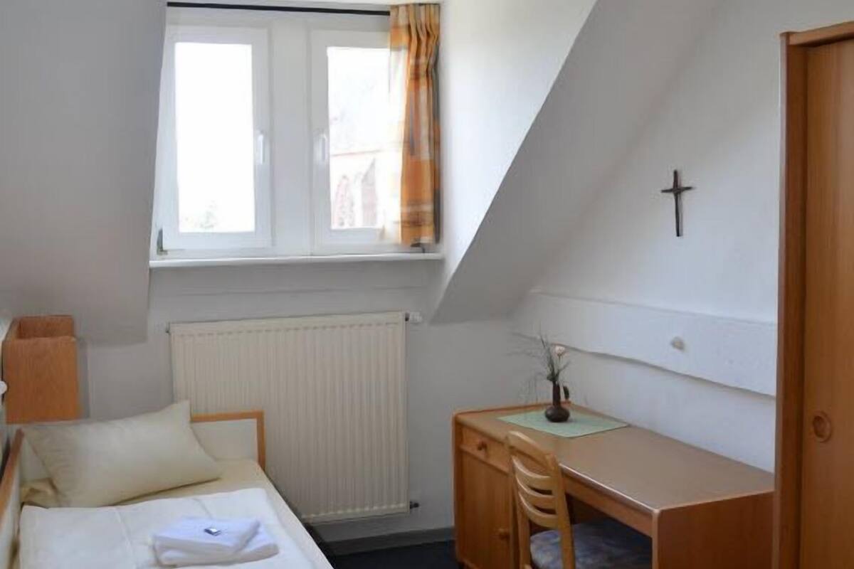 A small, simple room with a bed, a desk, and a chair. There is a window with curtains, a radiator, and a cross on the wall.