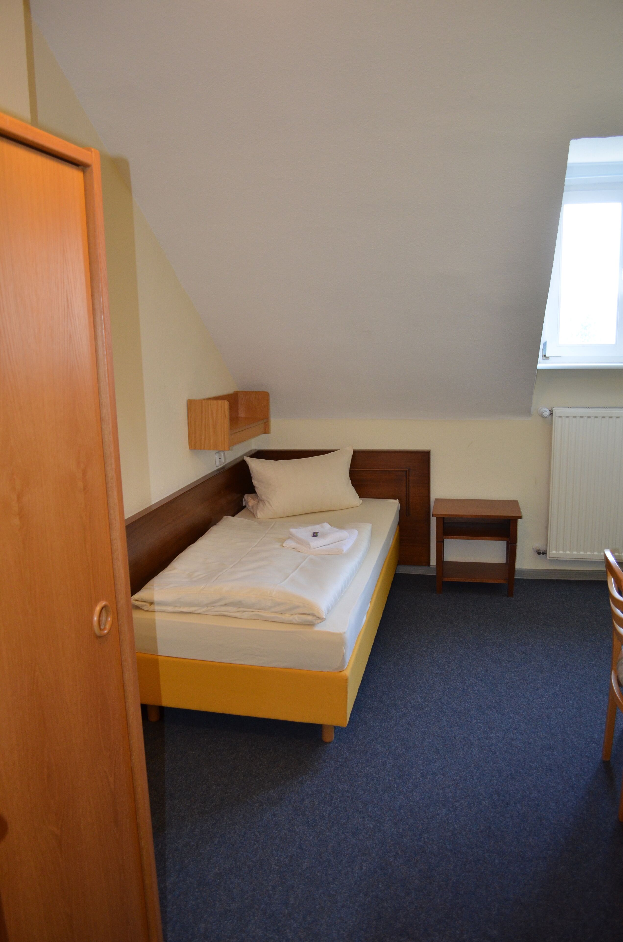 A bedroom with a single bed, a bedside table, a small wooden nightstand, a radiator, and a window with natural light.