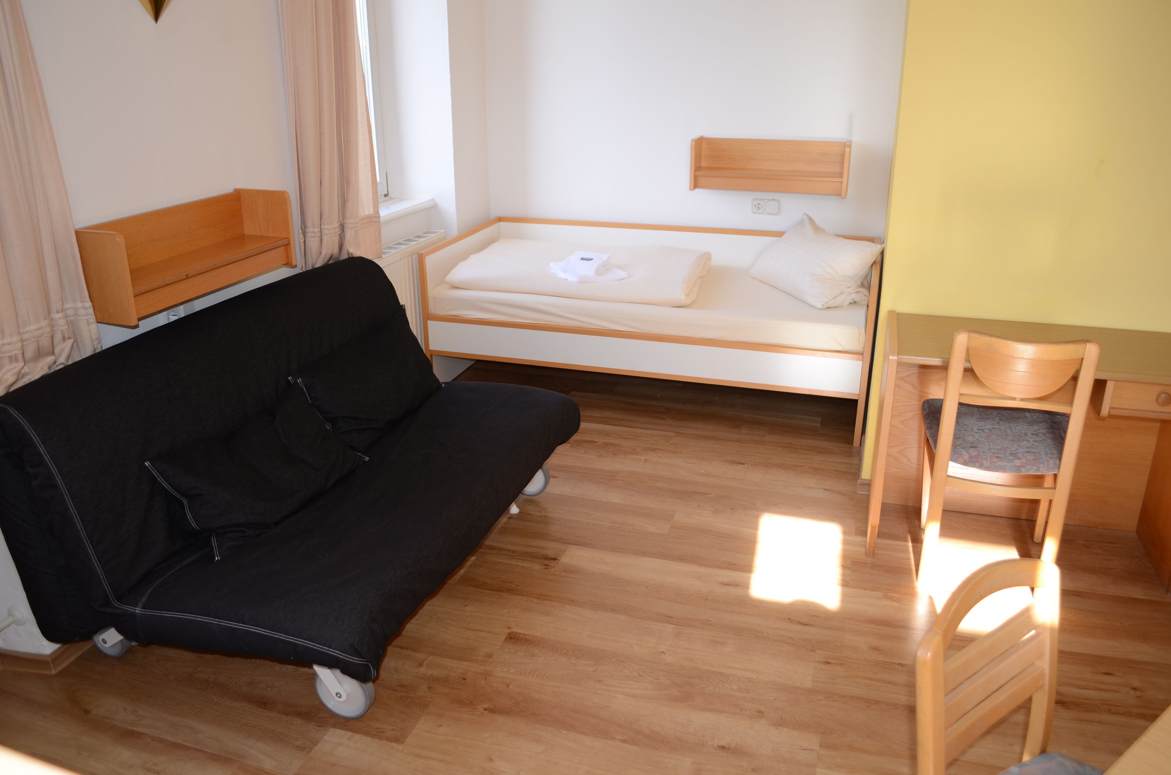 A compact room with a single bed, a small desk with a chair, and a sofa.