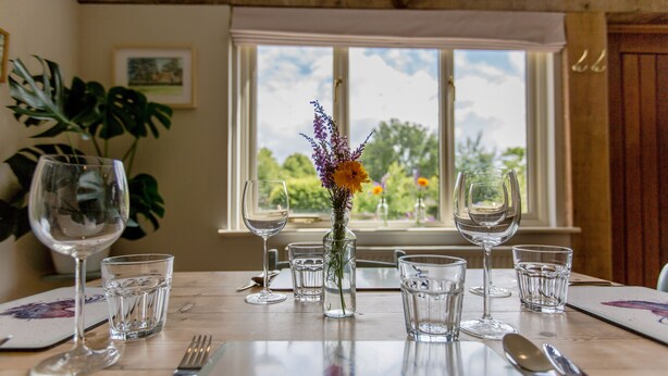 A dining table set with wine glasses, water glasses, and cutlery, with a vase of flowers in the center.