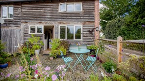 A rustic wooden house with a garden patio featuring a table and chairs, surrounded by potted plants and flowers.