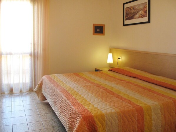 A hotel room with a bed, a nightstand, a lamp, a framed picture on the wall, and a window with curtains.