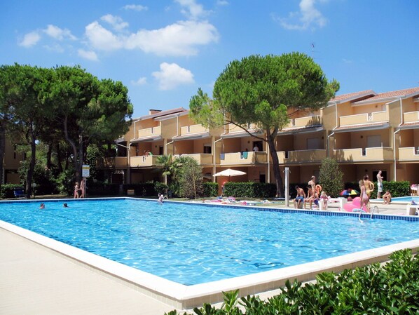 A swimming pool surrounded by multi-story buildings with balconies, trees, and a clear sky.