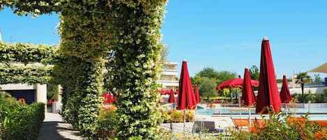 A pathway lined with red umbrellas leading to a pool area, surrounded by lush greenery and a clear blue sky.